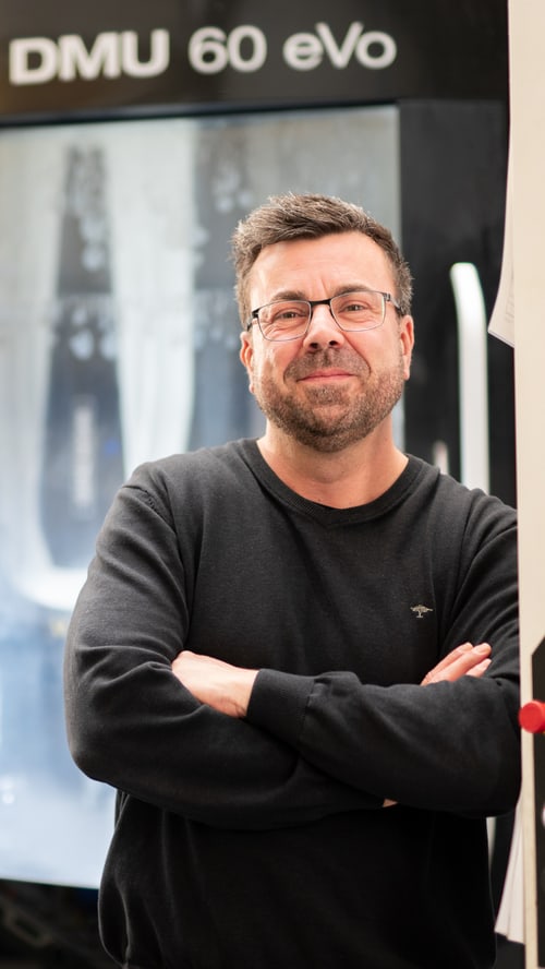 Mitarbeiter mit Brille und verschränkten Armen vor einer CNC-Fräsmaschine.